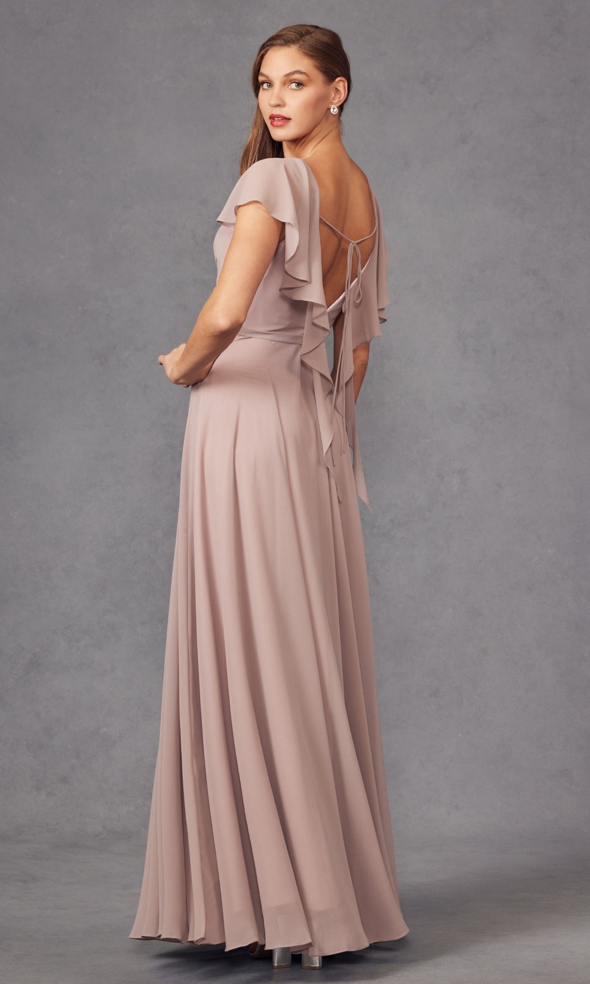 A woman in the Flutter-Sleeve Long Formal Evening Gown 261, featuring mauve fabric, flutter sleeves, and an open back with bow ties, stands against a gray background and glances over her shoulder.